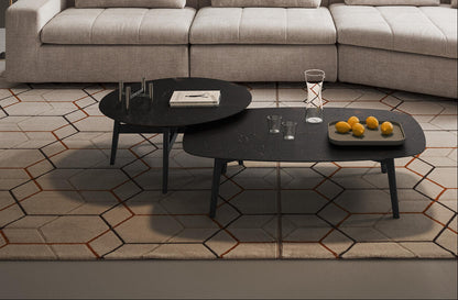 Two modern black coffee tables with tapered legs, styled in a living room setting on a geometric patterned rug in front of a light beige sectional sofa.