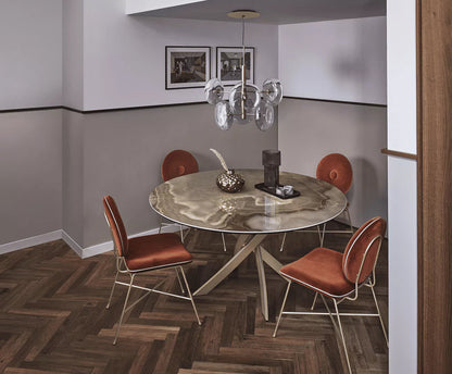 A modern dining room setup featuring a round marble-top table with a unique shaped  base, surrounded by four copper-brown upholstered chairs with gold metal frames. The room has a gray and white two-tone wall, a glass chandelier, and dark wood herringbone flooring.