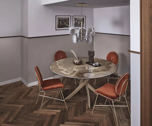 A modern dining room setup featuring a round marble-top table with a unique shaped  base, surrounded by four copper-brown upholstered chairs with gold metal frames. The room has a gray and white two-tone wall, a glass chandelier, and dark wood herringbone flooring.