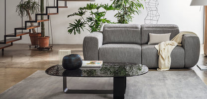 A modern gray upholstered sofa in a bright, contemporary living room, styled with a cream throw blanket, a black marble-top coffee table, and indoor plants.